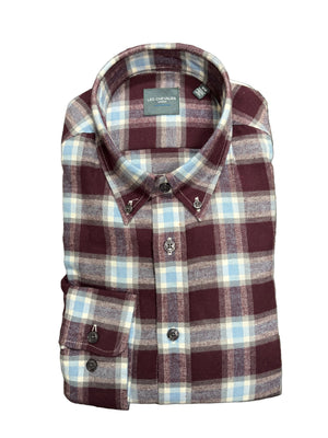 A folded long-sleeve plaid button-down shirt by Lee Chevalier, featuring a burgundy, light blue, and white check pattern with silver-tone skull-shaped buttons, laid flat on a textured green carpet.