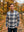 A man wearing a blue and beige plaid button-down shirt stands outdoors on a leaf-covered path in an autumn park.