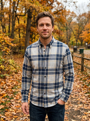 A man wearing a blue and beige plaid button-down shirt stands outdoors on a leaf-covered path in an autumn park.