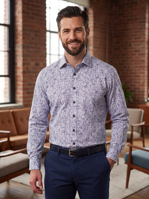 A man wearing a light purple and white patterned long-sleeve button-down shirt with navy blue trousers, standing in a modern office lounge with brick walls and large windows.
