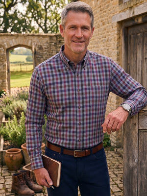 A smiling man stands outdoors in front of a rustic stone building, wearing a long-sleeve button-down plaid shirt in purple, red, and blue tones, navy trousers, and a brown belt. He is holding a brown leather notebook or journal.