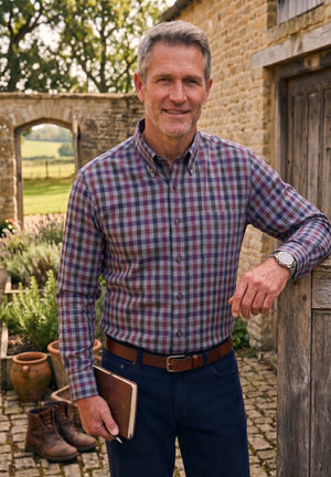 A smiling man stands outdoors in front of a rustic stone building, wearing a long-sleeve button-down plaid shirt in purple, red, and blue tones, navy trousers, and a brown belt. He is holding a brown leather notebook or journal.