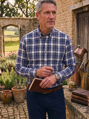 A man standing outdoors in front of a stone wall and rustic building, wearing a blue and white plaid button-down shirt, navy trousers, and holding a brown leather notebook.