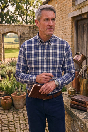 A man standing outdoors in front of a stone wall and rustic building, wearing a blue and white plaid button-down shirt, navy trousers, and holding a brown leather notebook.