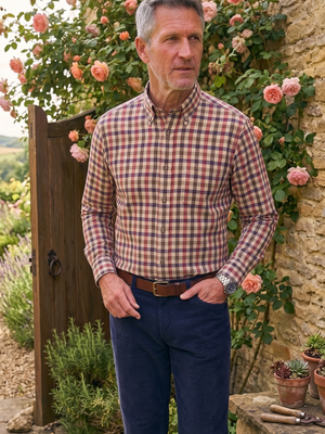 A man wearing a red and blue gingham long-sleeve button-down shirt and navy blue trousers stands in a rustic garden setting with stone walls, climbing roses, and potted plants.