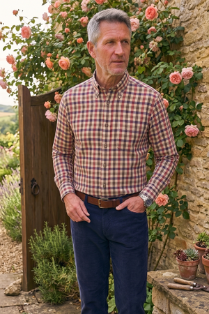 A man wearing a red and blue gingham long-sleeve button-down shirt and navy blue trousers stands in a rustic garden setting with stone walls, climbing roses, and potted plants.