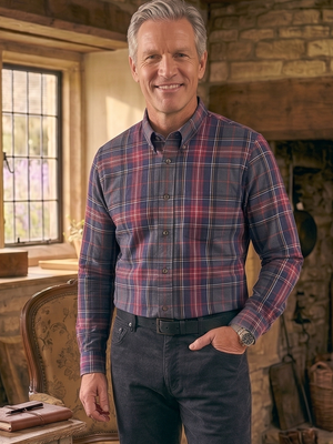 A smiling man with gray hair stands in a rustic stone room wearing a red and blue plaid button-down shirt, dark trousers, and brown boots. A wooden table with books and a glass lamp is on the left, and a stone fireplace is in the background.