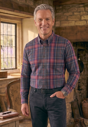 A smiling man with gray hair stands in a rustic stone room wearing a red and blue plaid button-down shirt, dark trousers, and brown boots. A wooden table with books and a glass lamp is on the left, and a stone fireplace is in the background.