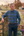 A man stands outdoors in a rural setting, leaning against a stone pillar next to a wooden gate. He is wearing a blue and green plaid button-down shirt and dark trousers.