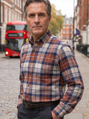 A man wearing a brown, blue, and cream plaid button-down shirt stands on a cobblestone street with a red double-decker bus in the background.
