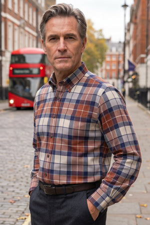 A man wearing a brown, blue, and cream plaid button-down shirt stands on a cobblestone street with a red double-decker bus in the background.