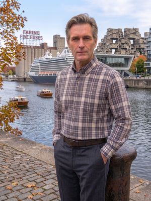 A man wearing a brown and beige plaid button-down shirt and dark trousers stands outdoors by a waterfront canal, with boats, a cruise ship, and urban buildings in the background.