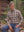 A man with gray hair and a beard sits on a wooden log by a lake, wearing a long-sleeve button-down plaid shirt in beige, brown, and navy blue, paired with blue jeans.
