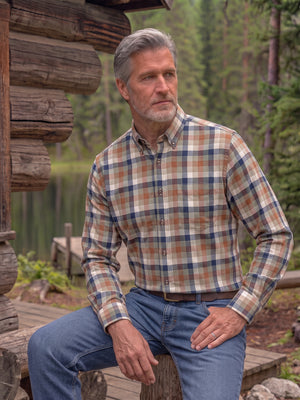 A man with gray hair and a beard sits on a wooden log by a lake, wearing a long-sleeve button-down plaid shirt in beige, brown, and navy blue, paired with blue jeans.