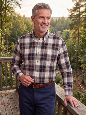 A smiling man wearing a long-sleeve plaid button-down shirt in beige, brown, and navy, paired with navy trousers and a brown belt, standing on a wooden deck with a forest background.