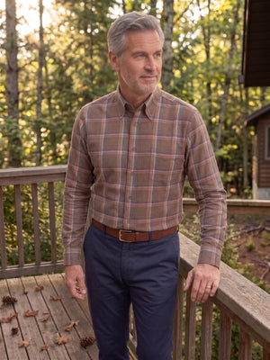 A man wearing a brown and beige plaid button-down shirt with navy blue pants, standing on a wooden deck with a forest background.