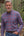 A man wearing a blue and red plaid button-down shirt stands outdoors in front of a log cabin and a forest lake.