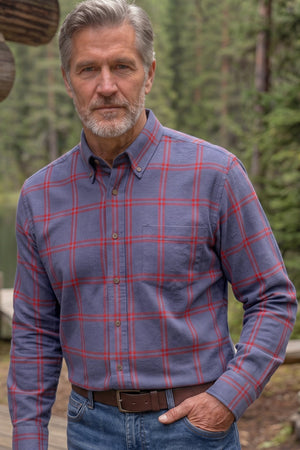 A man wearing a blue and red plaid button-down shirt stands outdoors in front of a log cabin and a forest lake.
