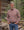 A man wearing a red, blue, and white plaid button-down shirt and blue jeans stands in front of a rustic log cabin with a forest background.