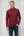 Man wearing a burgundy long-sleeve shirt and dark jeans standing against a light wooden panel background.