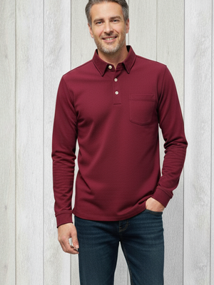 Man wearing a burgundy long-sleeve shirt and dark jeans standing against a light wooden panel background.