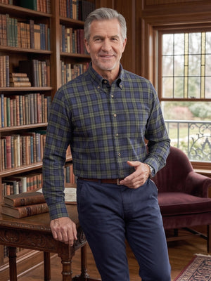 A man wearing a blue and green plaid button-down shirt and navy trousers stands in a classic library with wooden bookshelves, leaning on a wooden table.