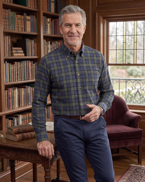 A man wearing a blue and green plaid button-down shirt and navy trousers stands in a classic library with wooden bookshelves, leaning on a wooden table.