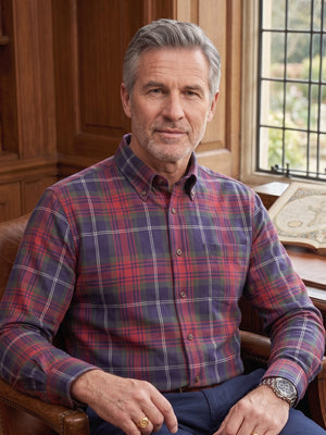 A man with gray hair sits in a leather armchair in a warm, wood-paneled study. He is wearing a long-sleeve button-down plaid shirt with red, blue, and purple tones, and dark trousers.