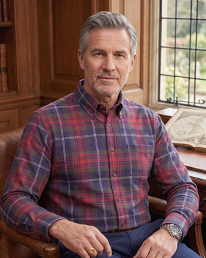 A man with gray hair sits in a leather armchair in a warm, wood-paneled study. He is wearing a long-sleeve button-down plaid shirt with red, blue, and purple tones, and dark trousers.